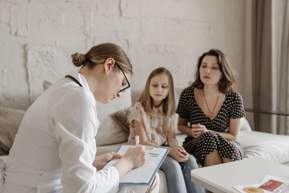 A family doctor consults with a mother and daughter in a warm, welcoming medical office setting.